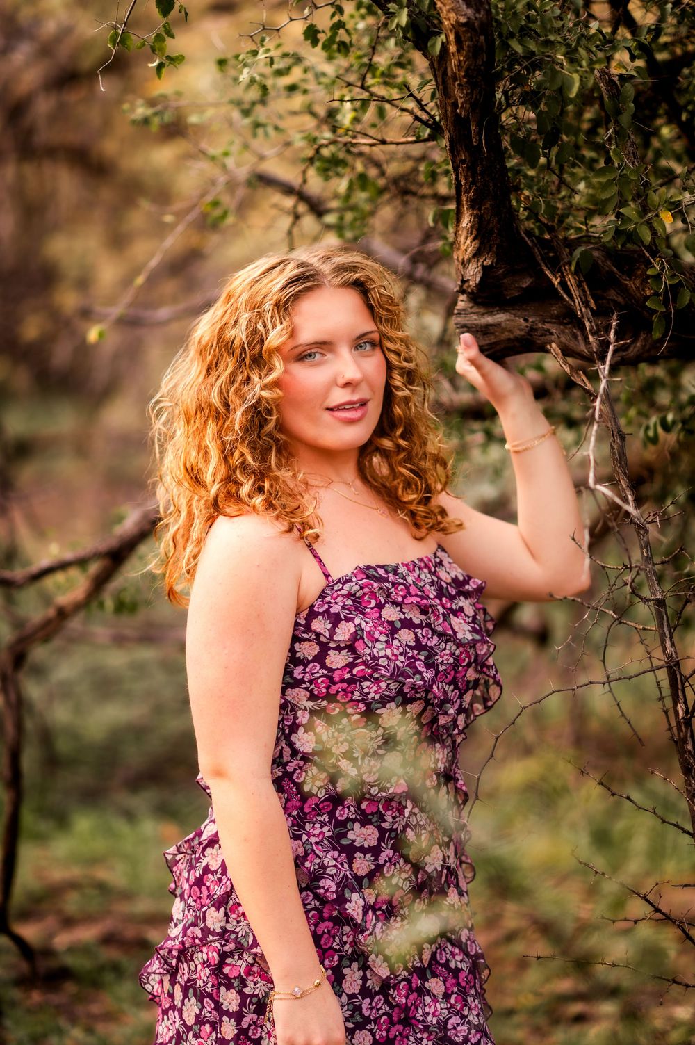 Young woman with curly red hair in floral dress poses by tree branch in natural outdoor setting.