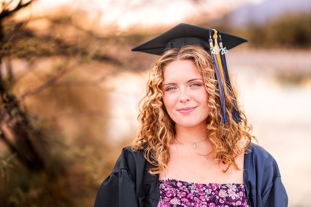 Young woman in graduation cap and gown smiling outdoors during golden hour in a desert setting.