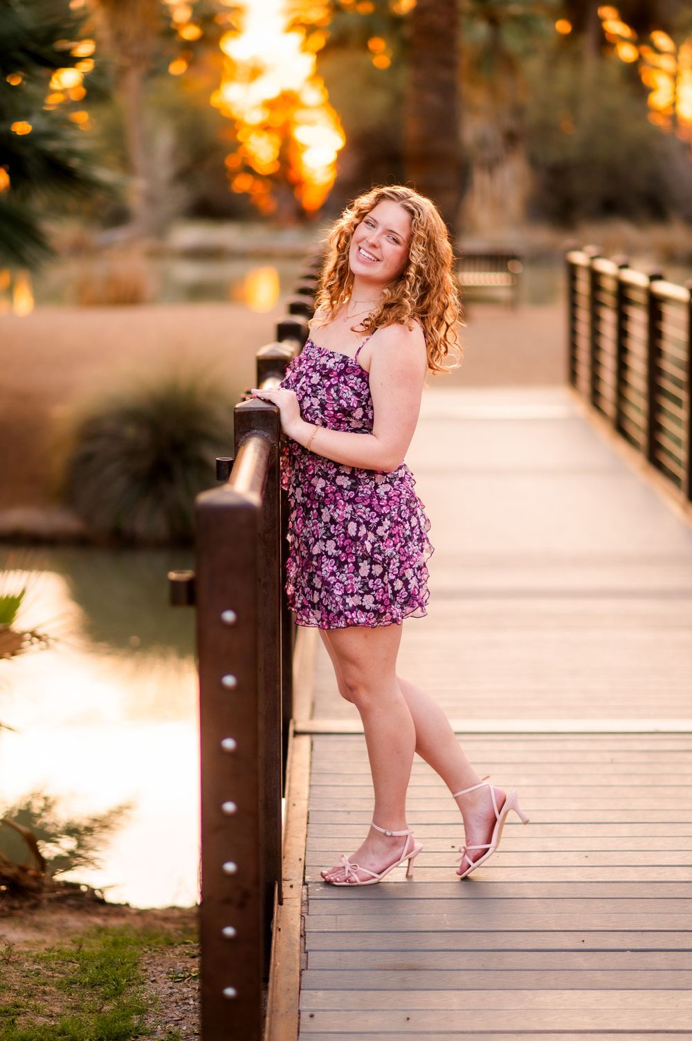 Young woman in floral purple dress and heels posing on wooden bridge at golden hour sunset.