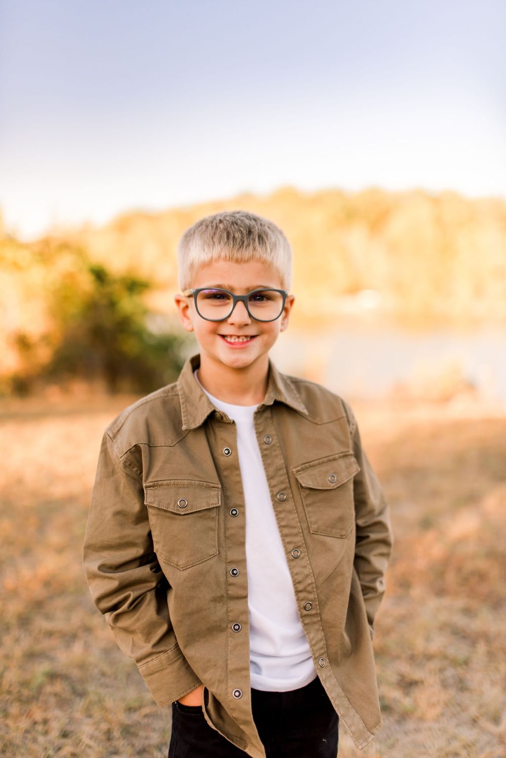 A child wearing glasses and brown jacket smiles outdoors during golden hour sunset in a natural setting.