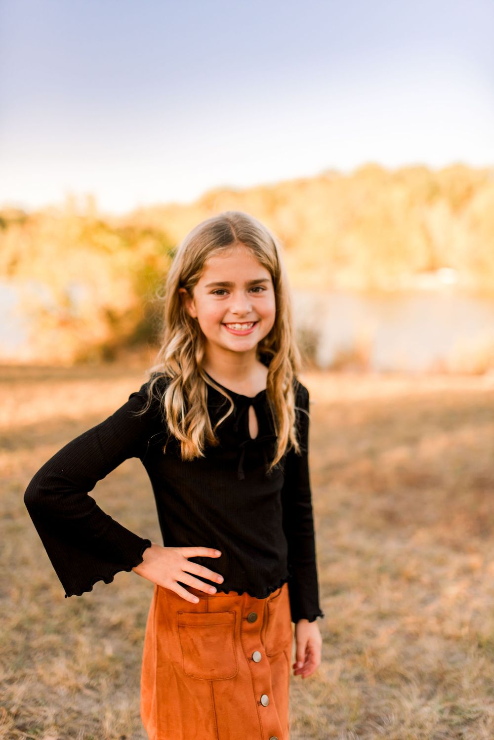 Young person in black top and brown corduroy skirt poses outdoors during golden hour sunset in autumn field.