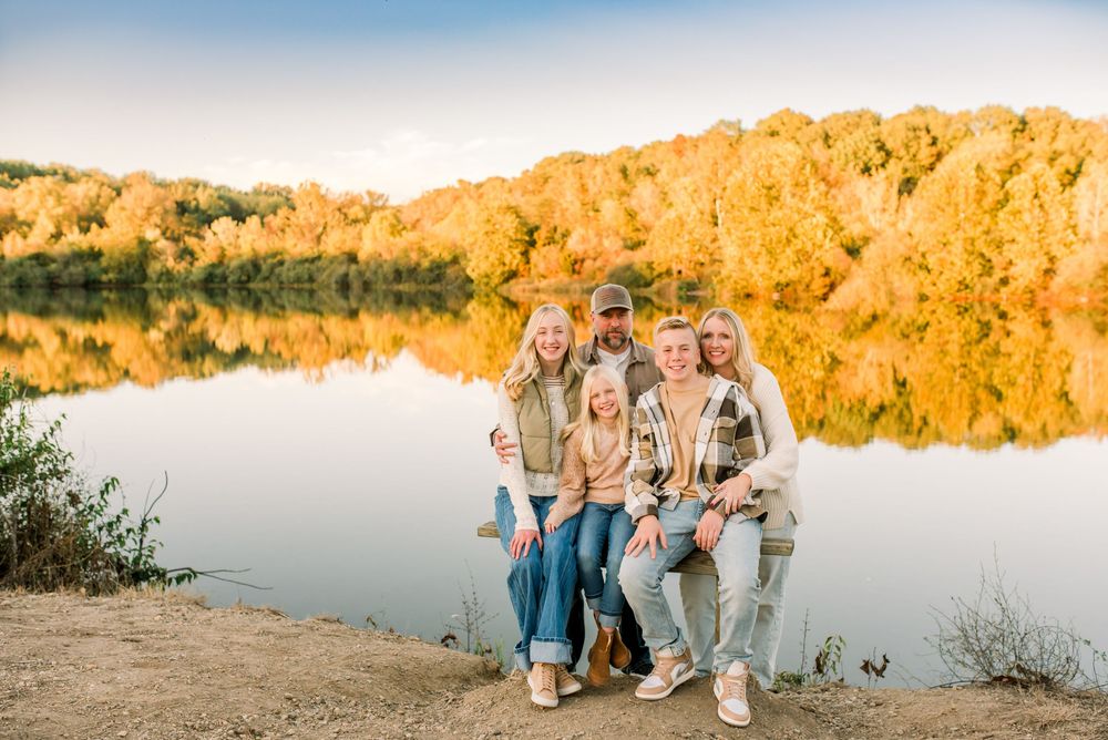 A group of people sit together on rocks near a serene lake at golden hour with autumn foliage in the background.