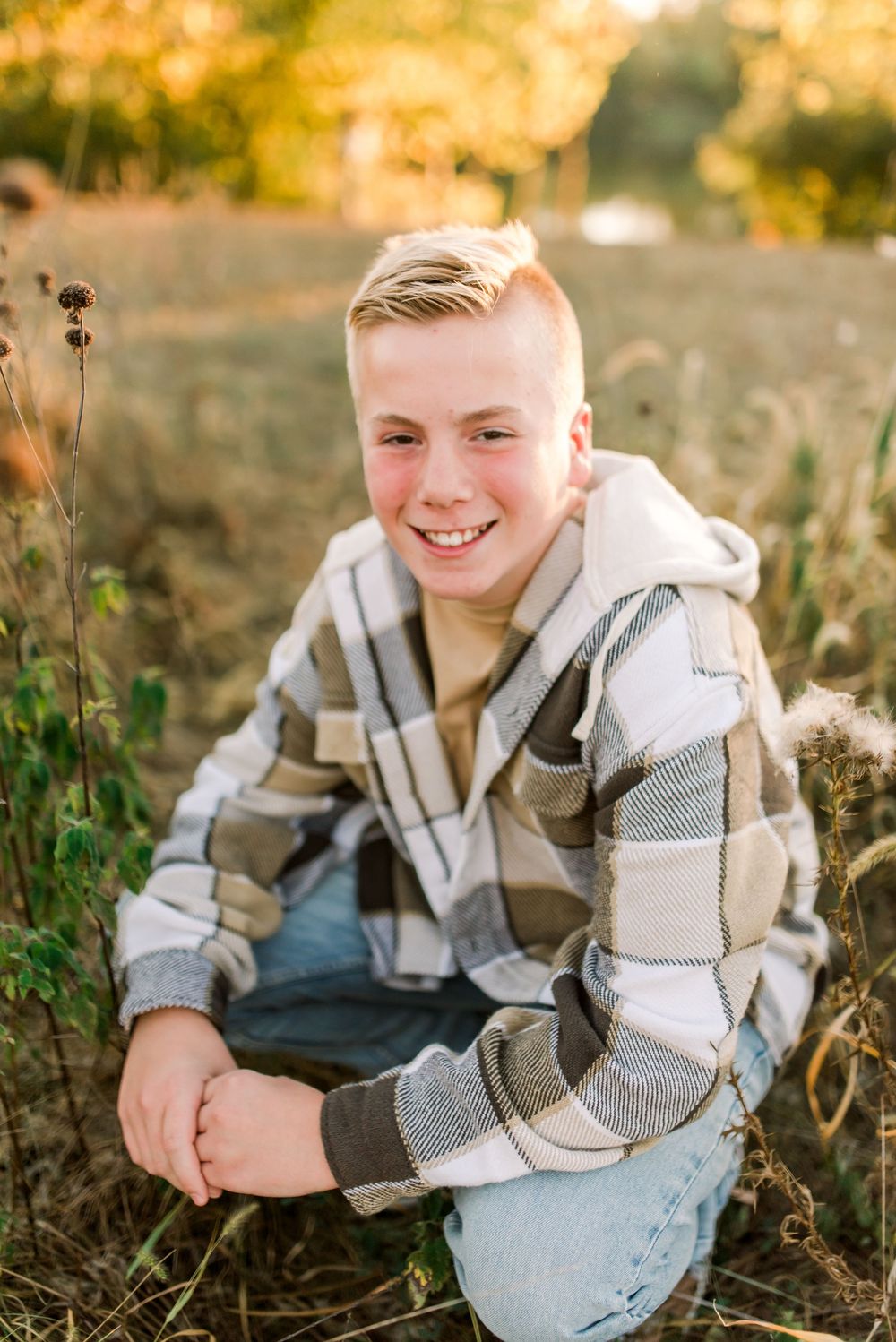 Smiling youth wearing a striped sweater sits in a sunlit field with dried plants during golden hour.