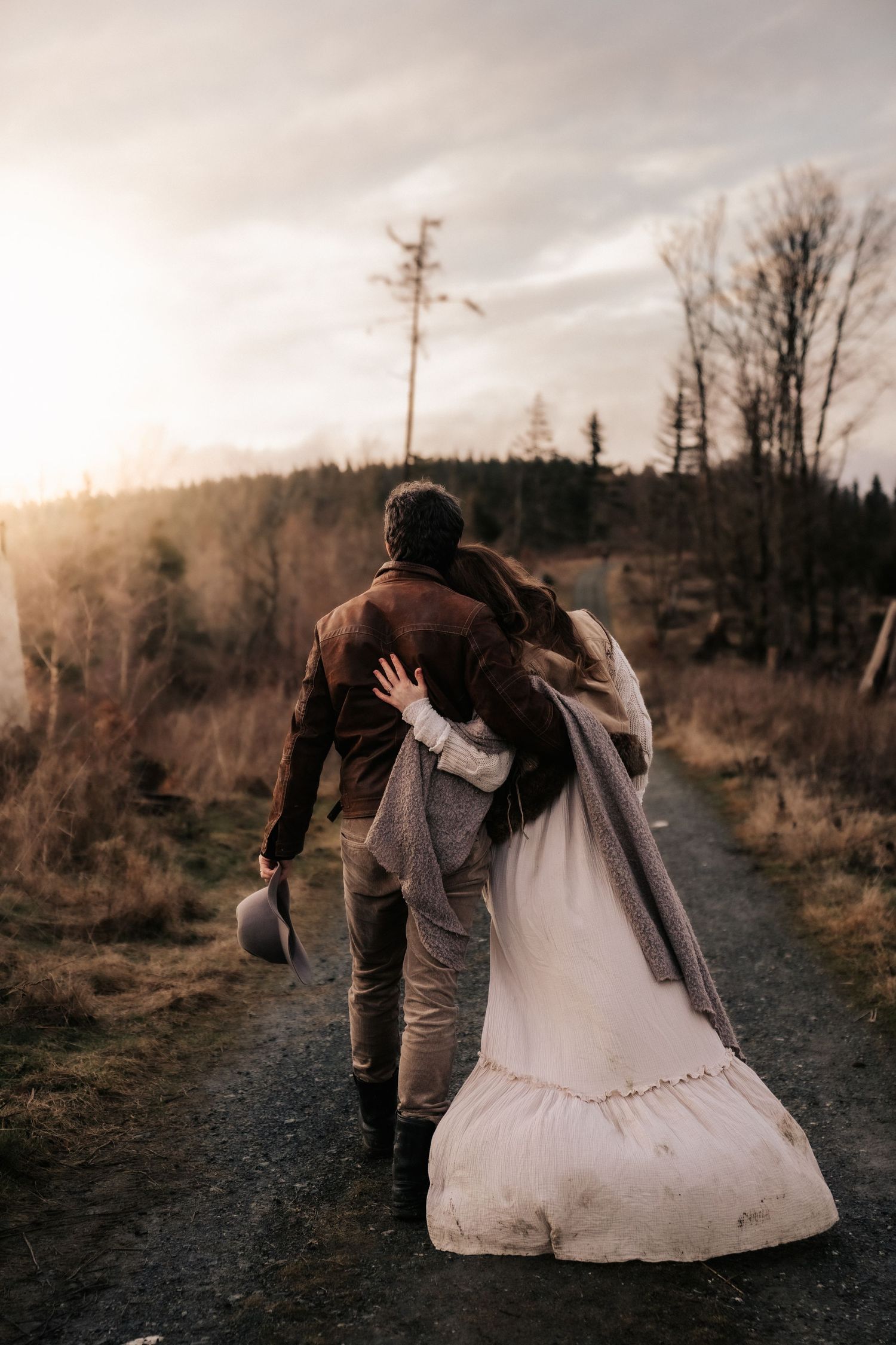 A couple in wedding attire walk together down a dirt path at sunset with trees and mountains in background.