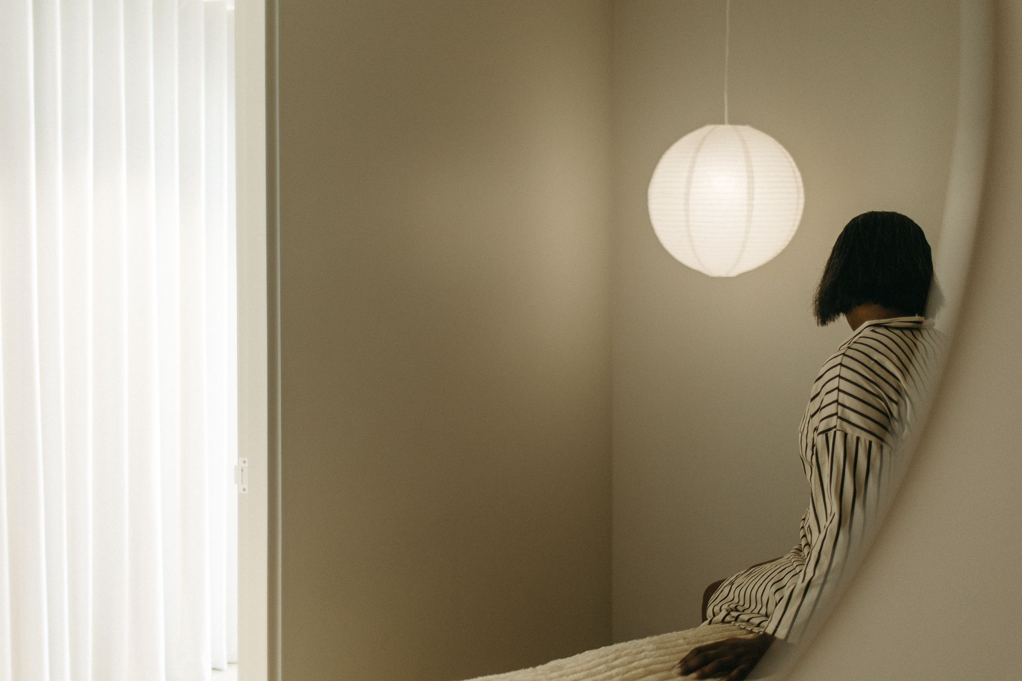 Person in striped shirt reflected in oval mirror beside glowing pendant light in minimalist beige interior.