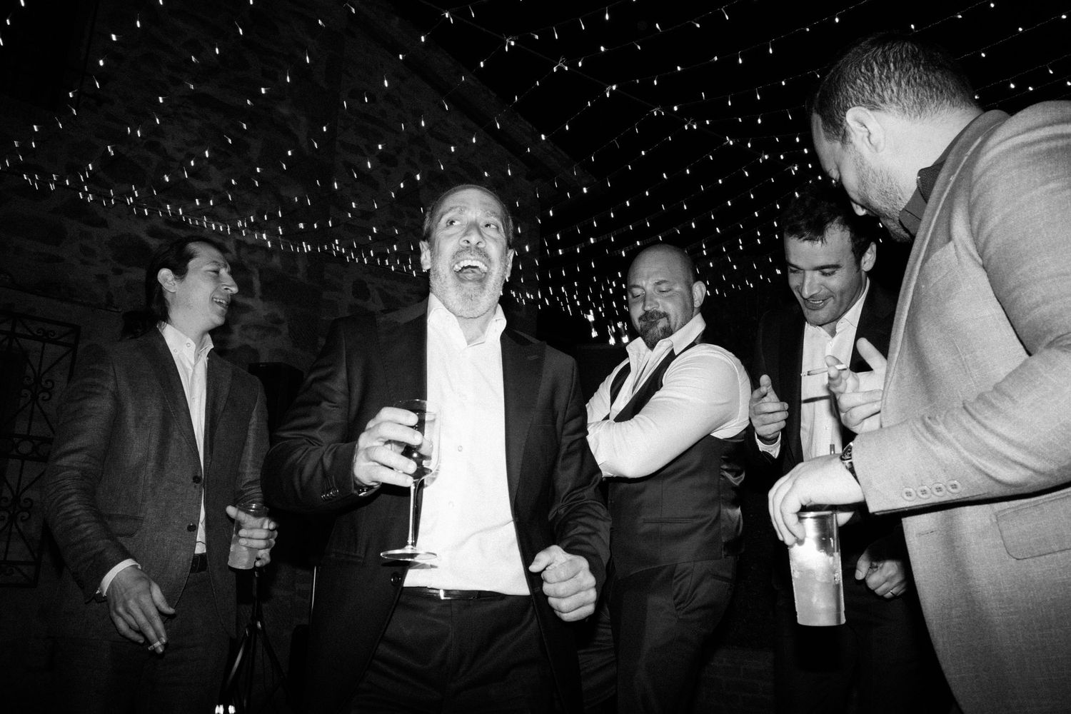 A black and white photo of people celebrating with drinks under string lights.