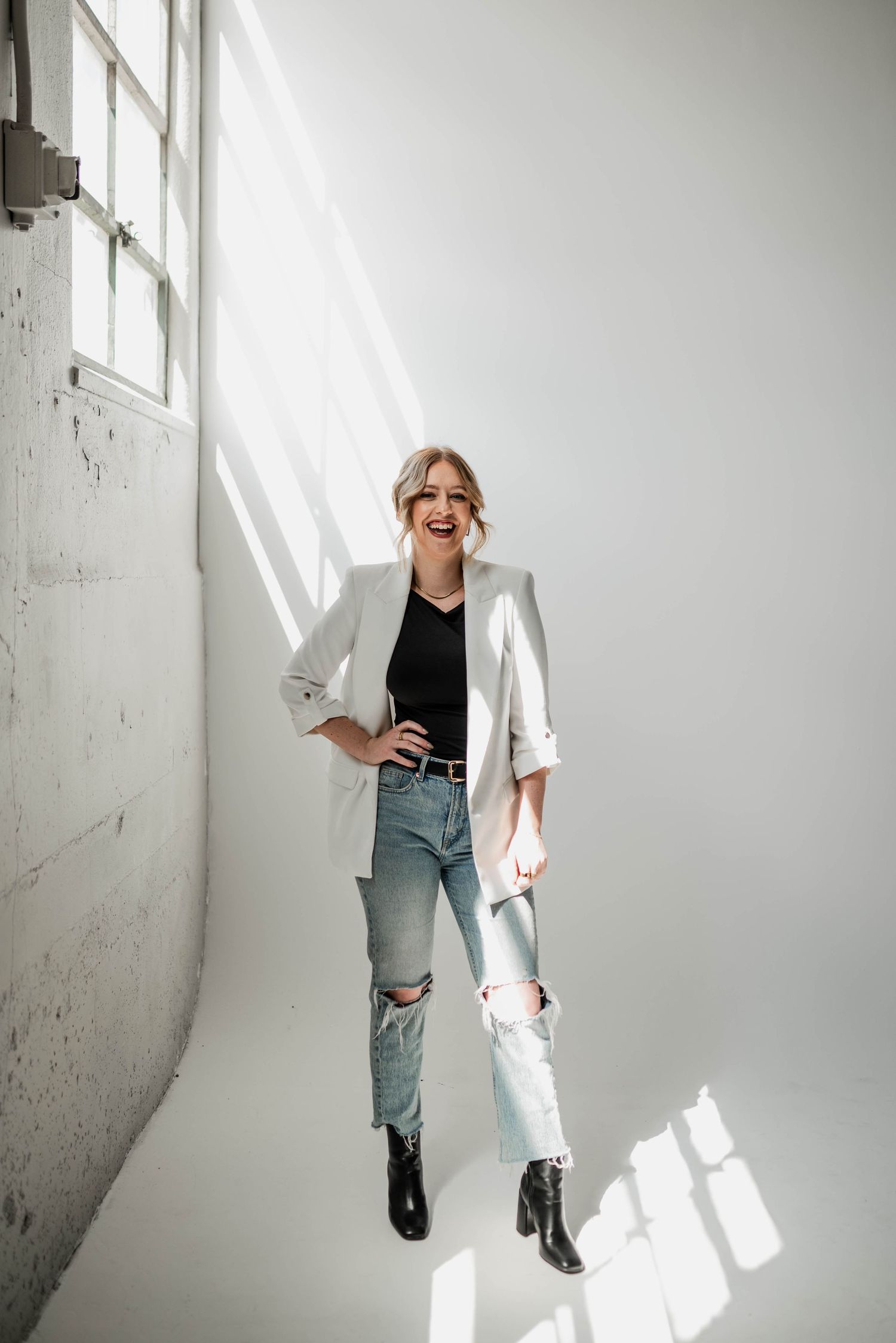 Stylish outfit featuring white blazer, ripped jeans and black boots in minimalist white studio setting.