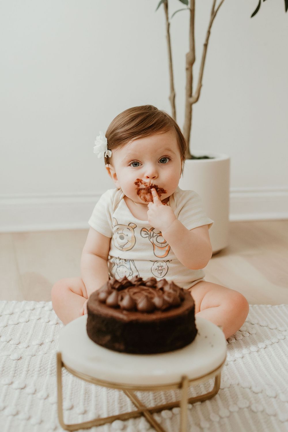 Baby in light colored outfit reaching for chocolate cake during cake smash photo session on white backdrop.
