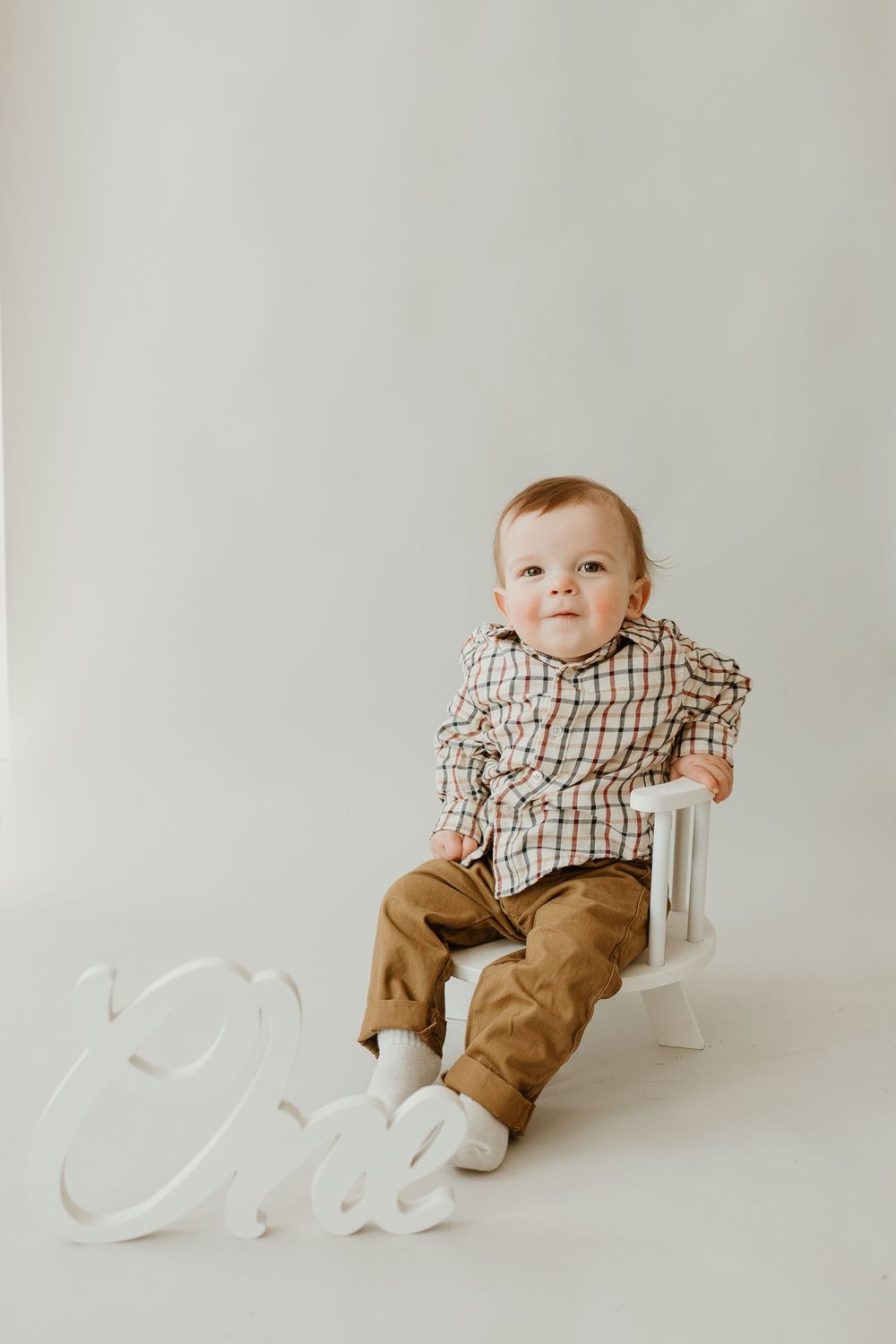 Baby in plaid shirt and brown pants sits on white chair against neutral background with 'One' text display.