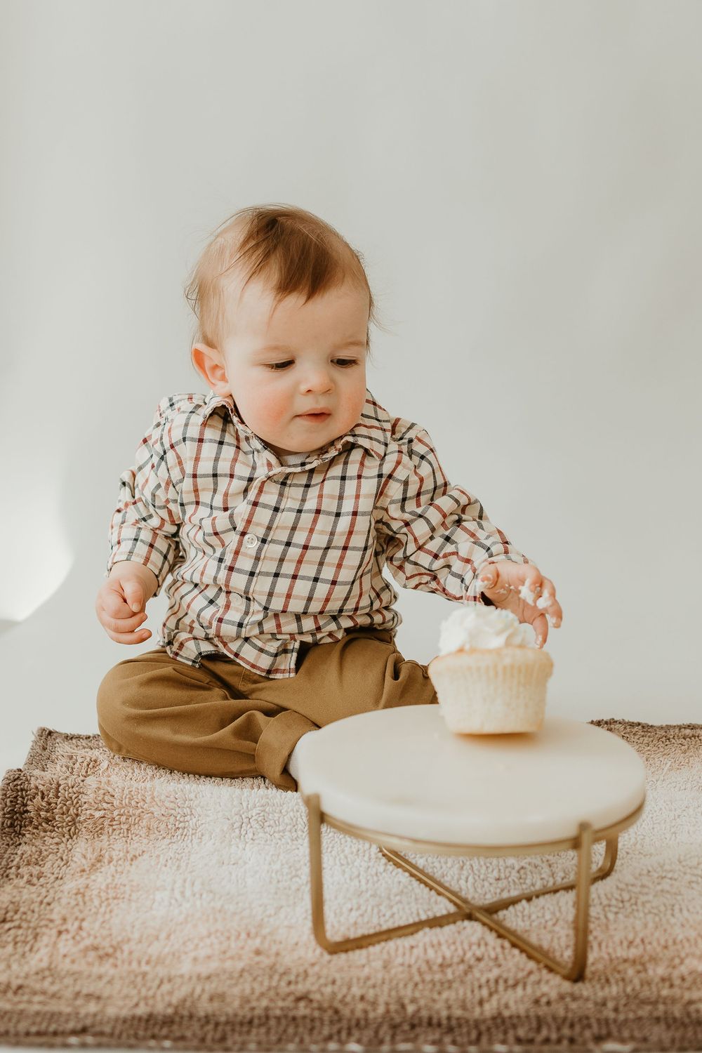 A baby in plaid shirt and khaki overalls reaches for a cupcake on a round white coffee table.