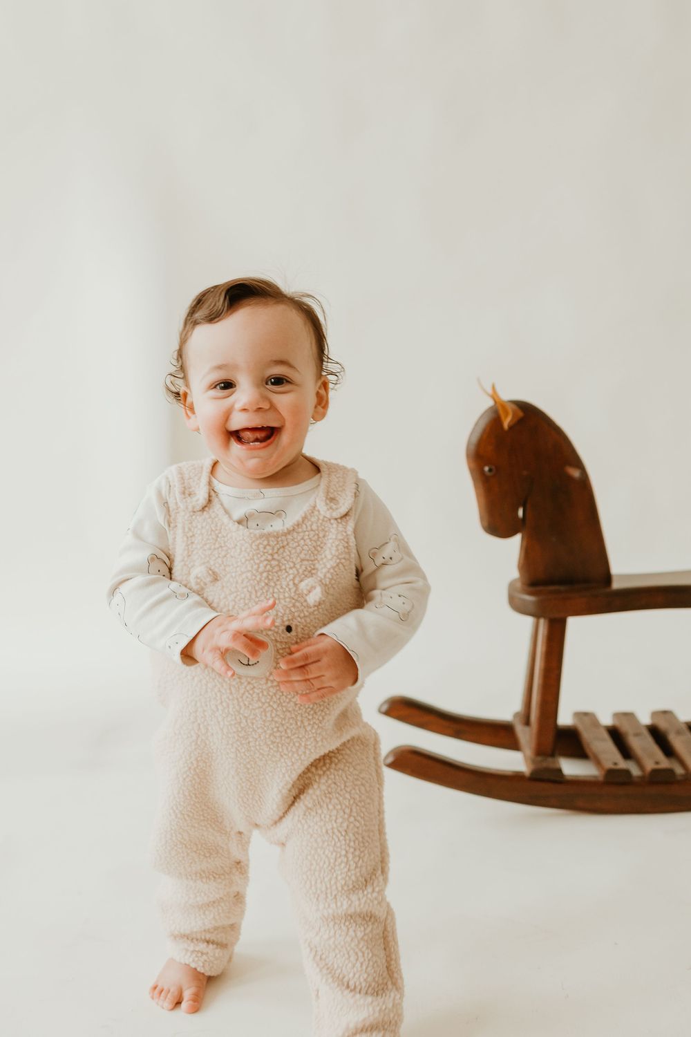 Happy baby in white knit romper laughs while standing next to wooden rocking horse toy against neutral background.