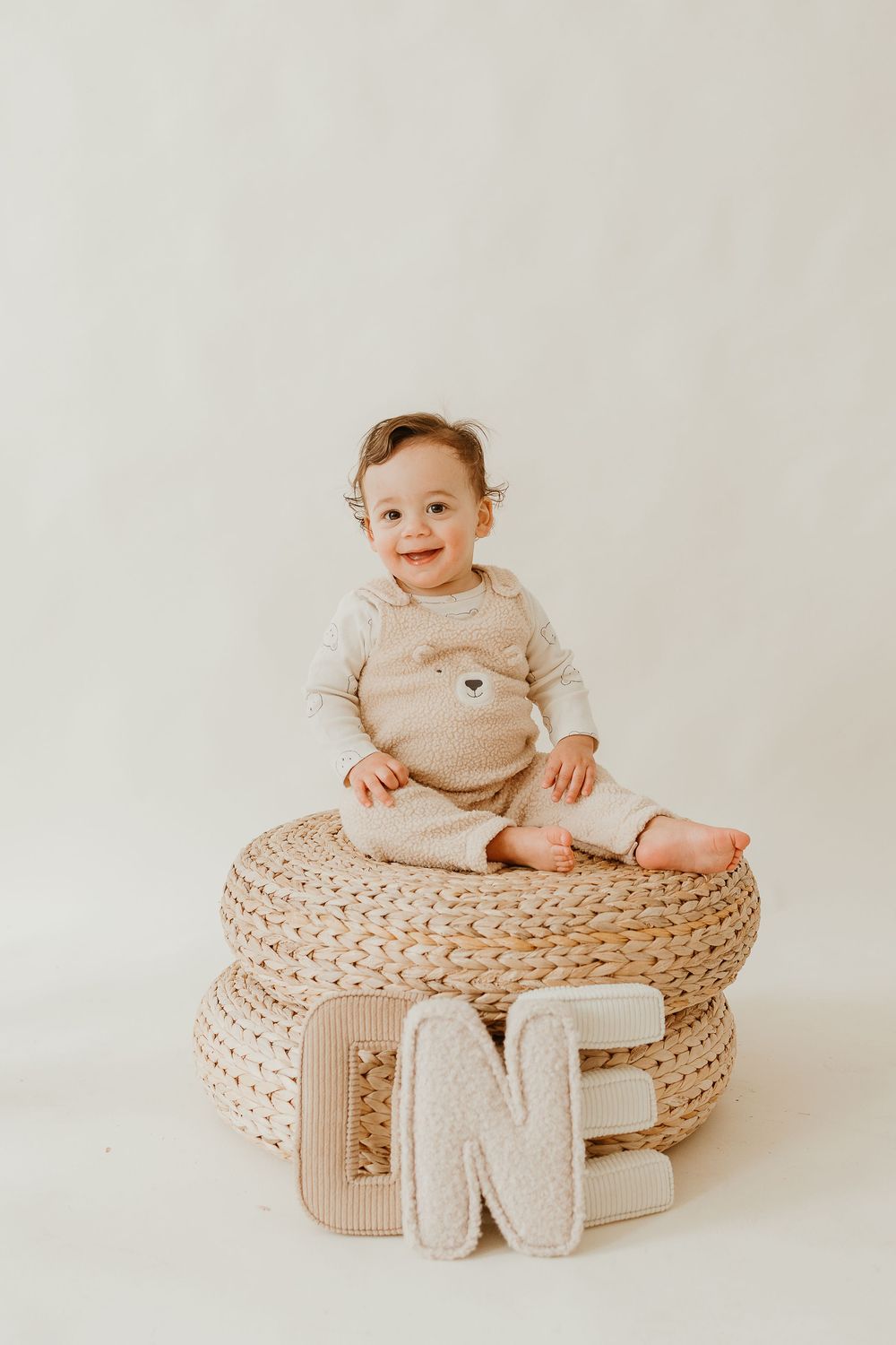 A baby in beige clothing sits atop a woven basket pouf with neutral-toned decorative elements in a minimalist setting.