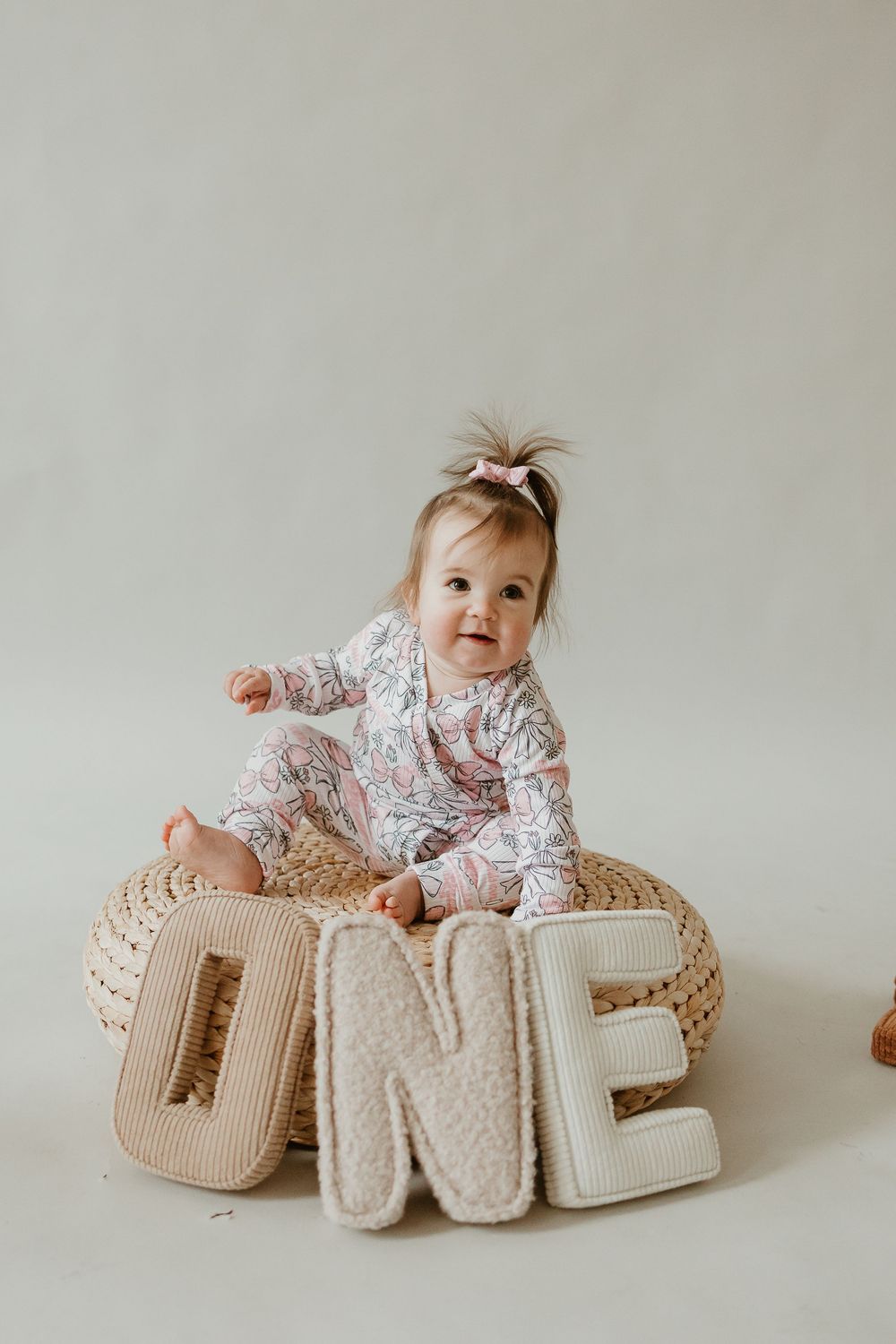 Baby in floral outfit lays beside decorative beige knit letters spelling 'ONE' against neutral background.