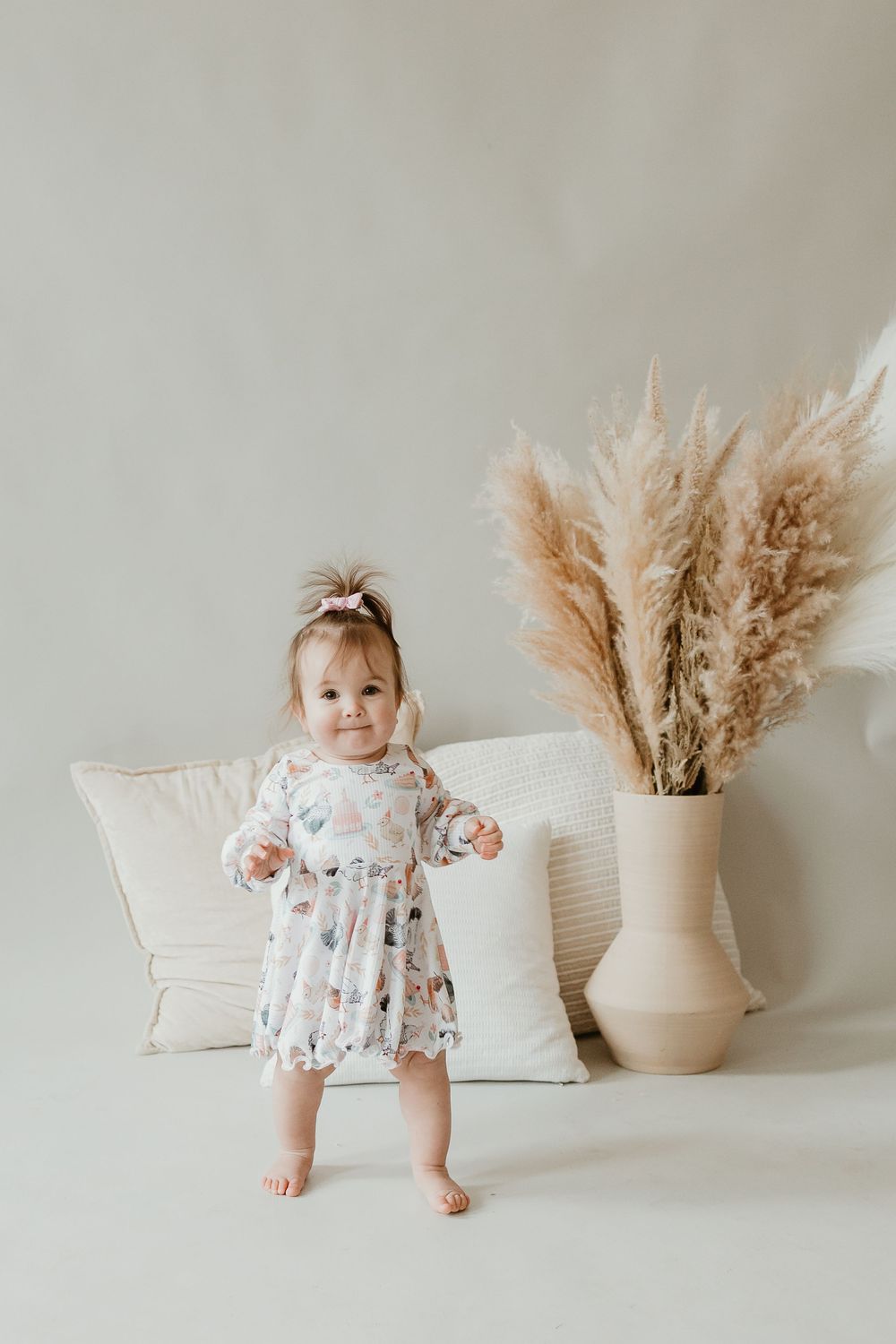 Minimalist nursery scene with floral dress, white cushions and decorative pampas grass in neutral beige vase.