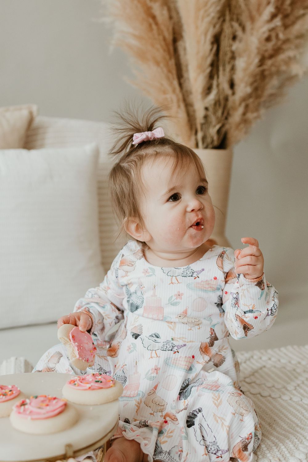 Baby in white patterned outfit enjoys pink frosted cake while sitting at table with pampas grass decor in background.