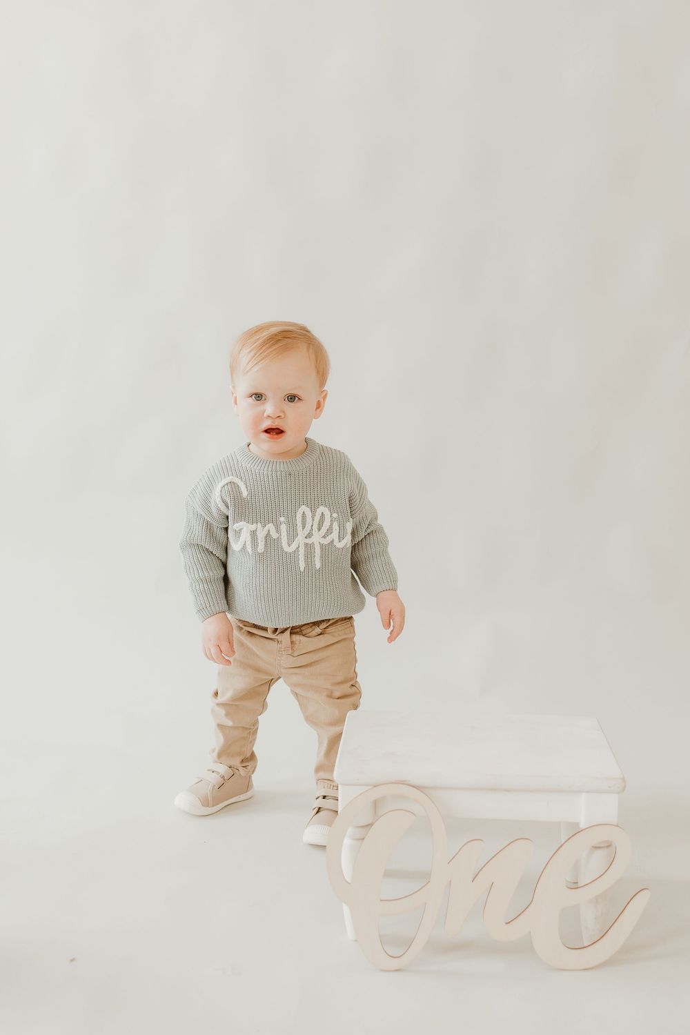 A toddler wearing a gray sweater and khaki pants stands next to a decorative 'one' sign against a white backdrop.