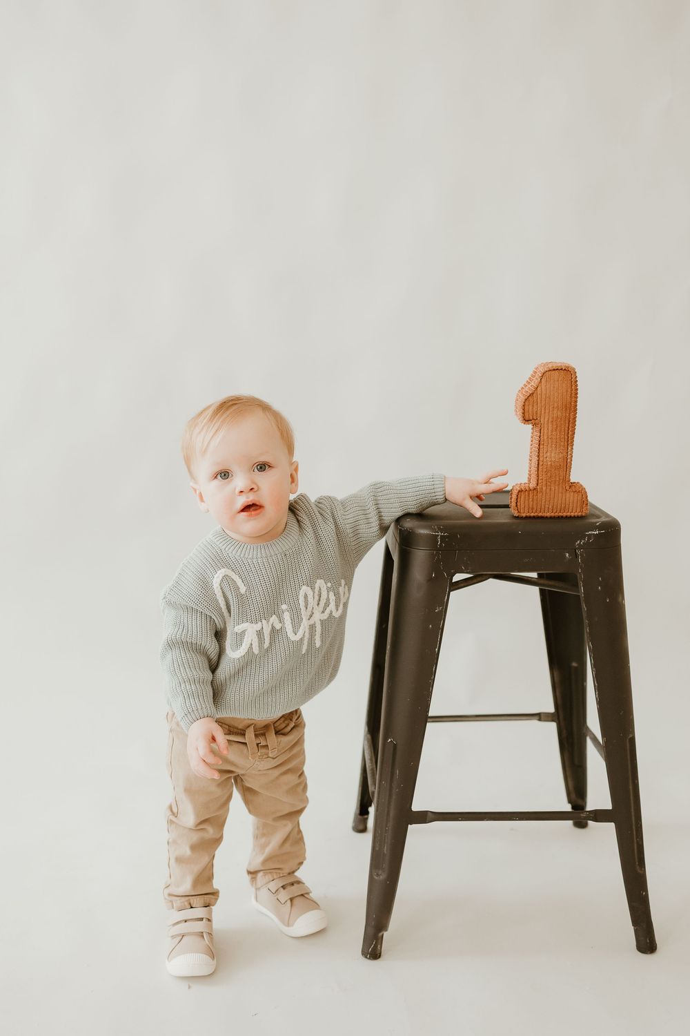 Baby in gray sweater and khaki pants explores dark metal stool against white background in progressive milestone photos.