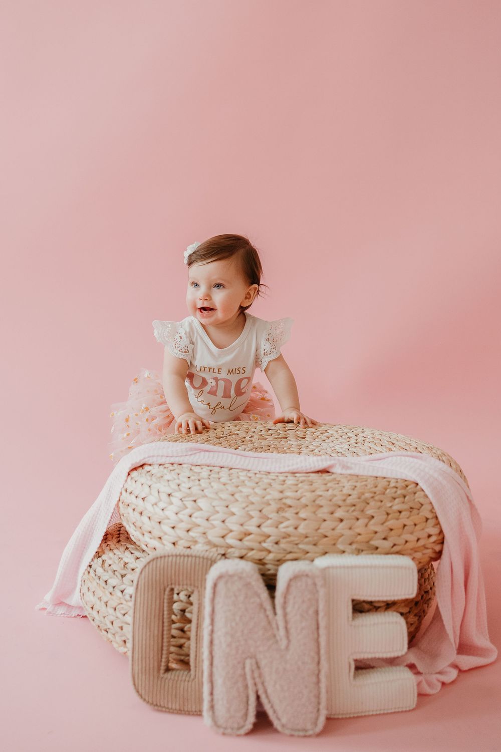 Baby in white dress crawls on large knitted pouf against pink backdrop in milestone photo session series.
