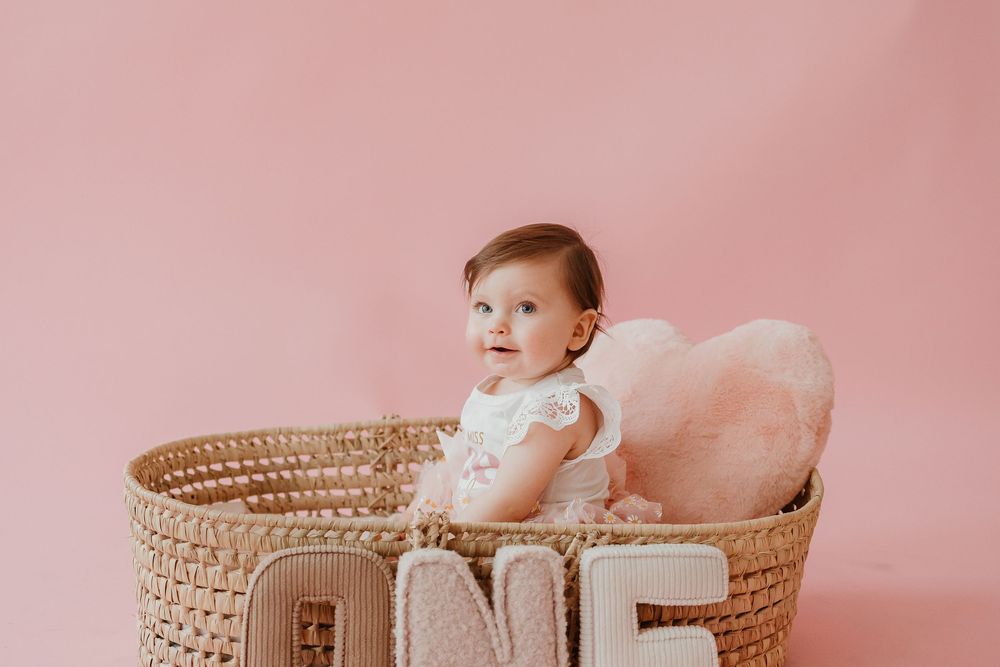 Baby in white dress sitting in woven basket with 'ONE' text against pink background.