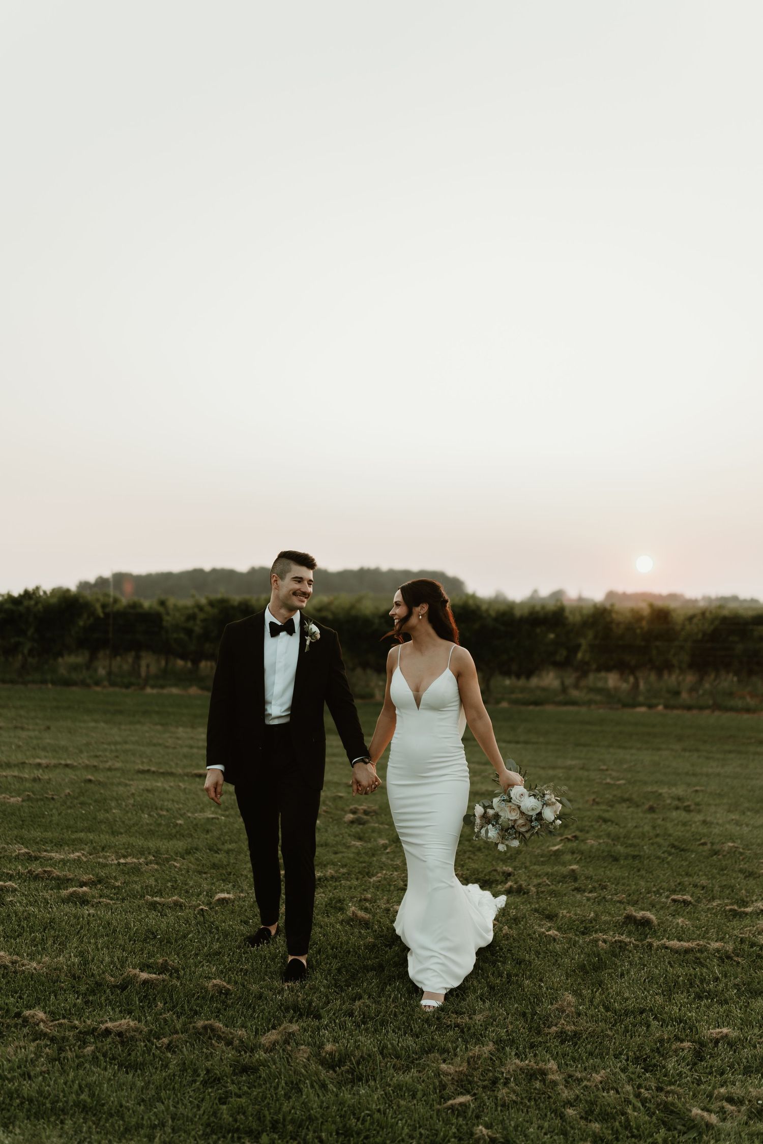 Bride and groom walk hand in hand through a field at sunset in formal wedding attire.