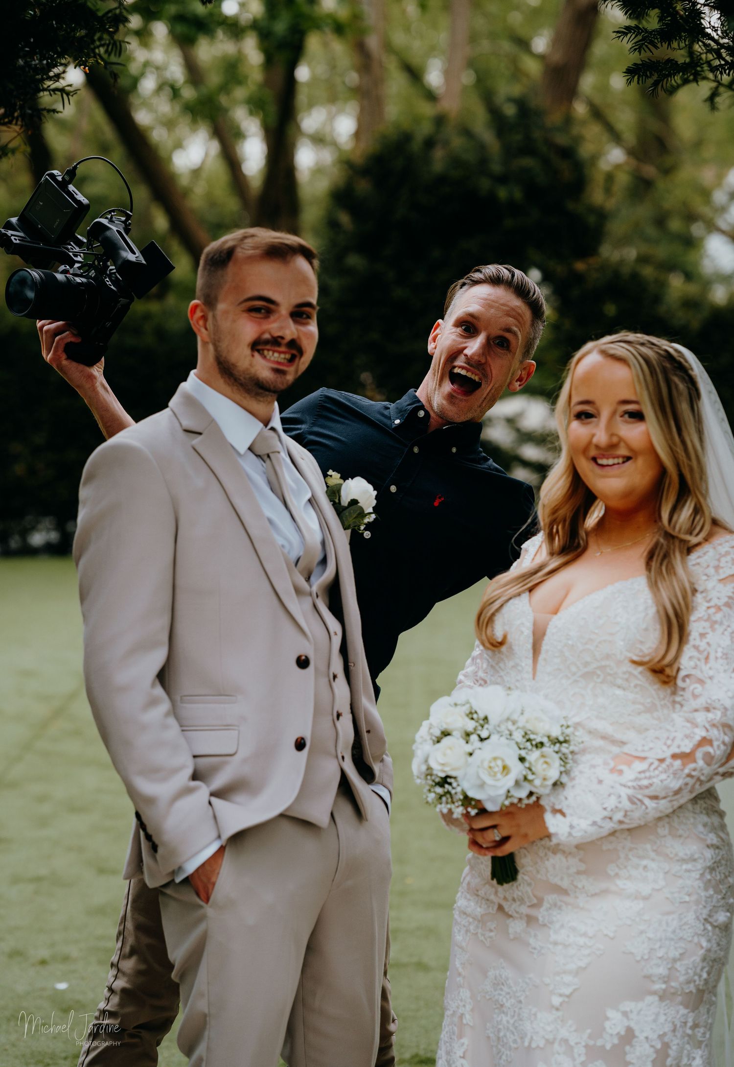Wedding videographer poses with newlyweds in an outdoor garden setting.