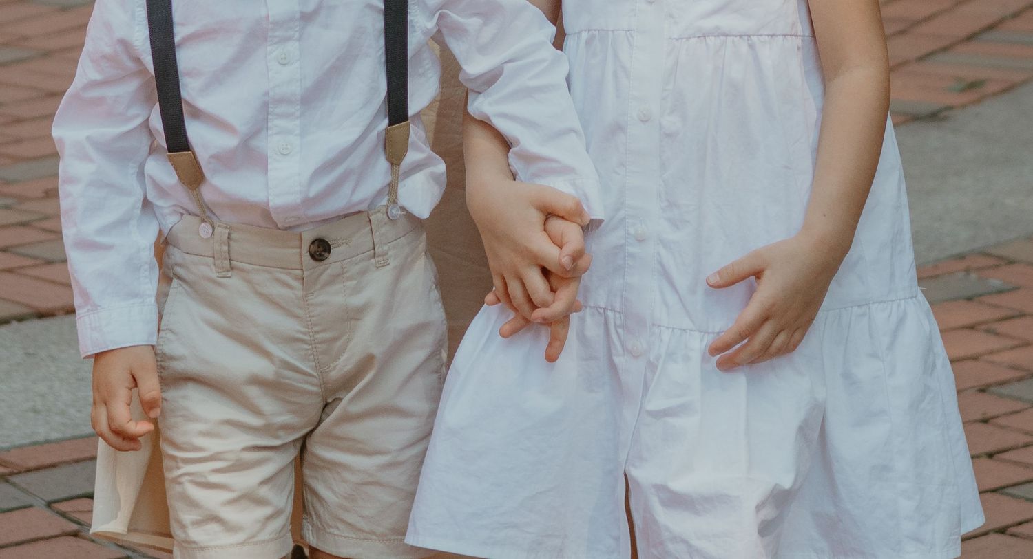 Children holding hands in white outfits with suspenders on brick pavement.