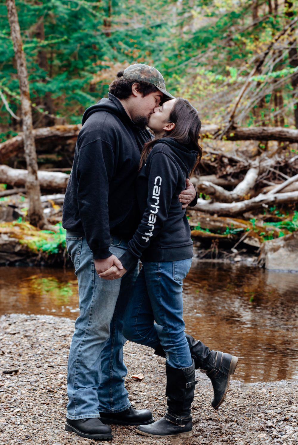 A romantic couple sharing a kiss by a creek in the woods wearing casual clothing and boots.