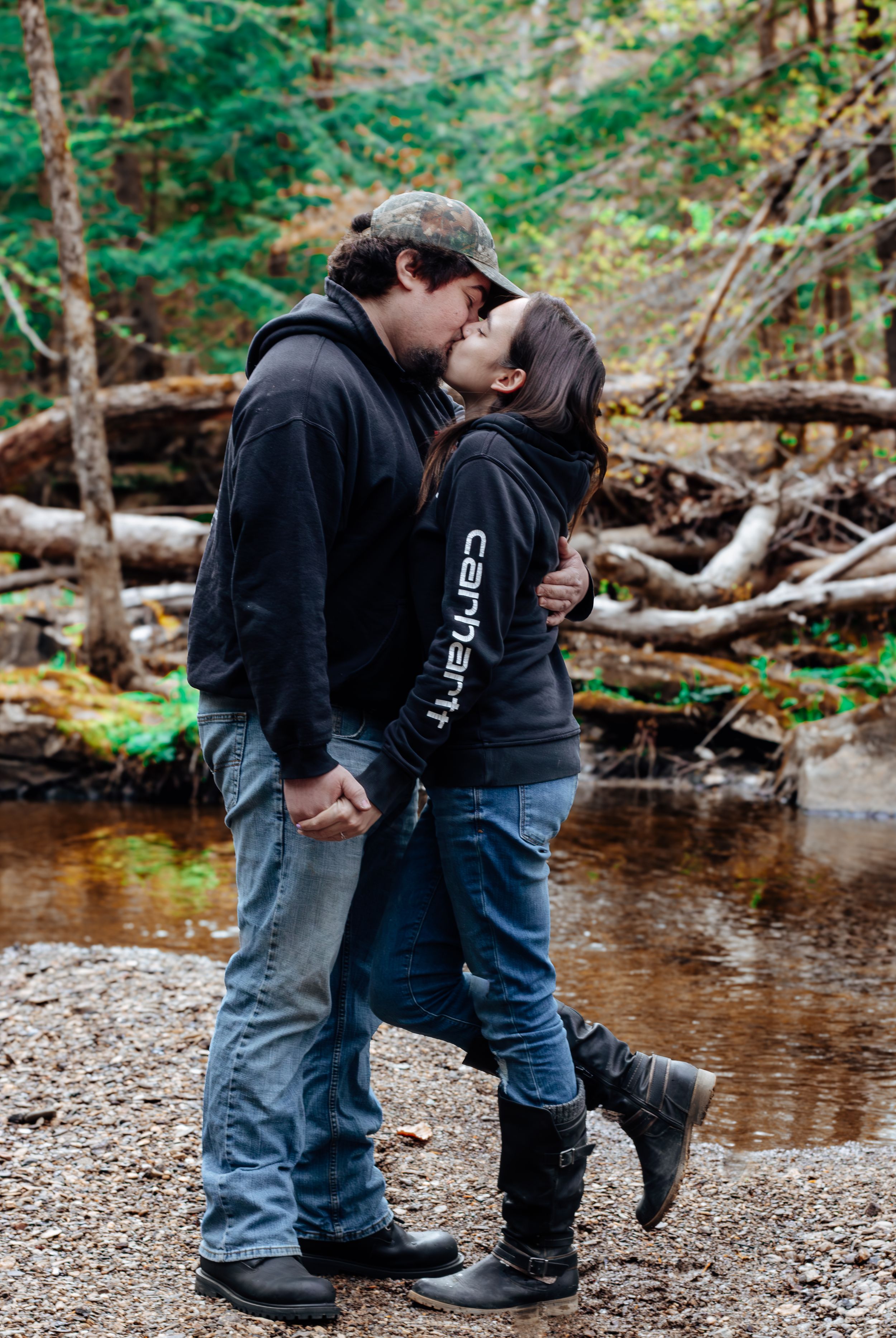 A romantic couple sharing a kiss by a creek in the woods wearing casual clothing and boots.