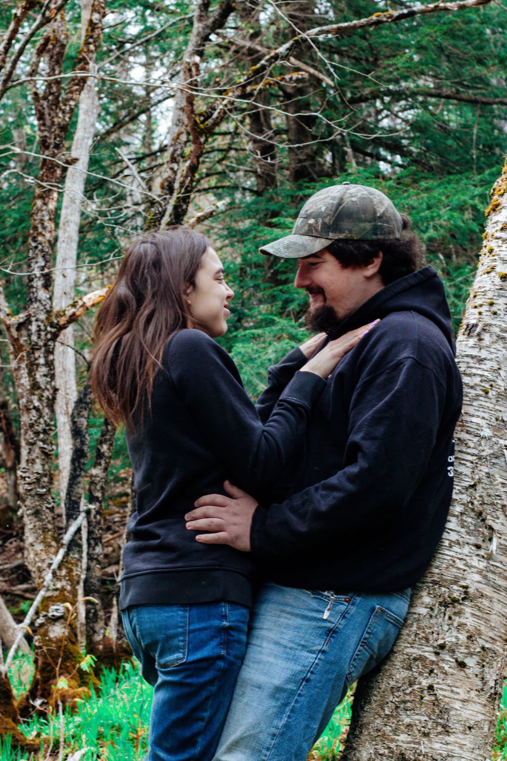 A couple in dark hoodies embracing against a birch tree in a lush forest setting.