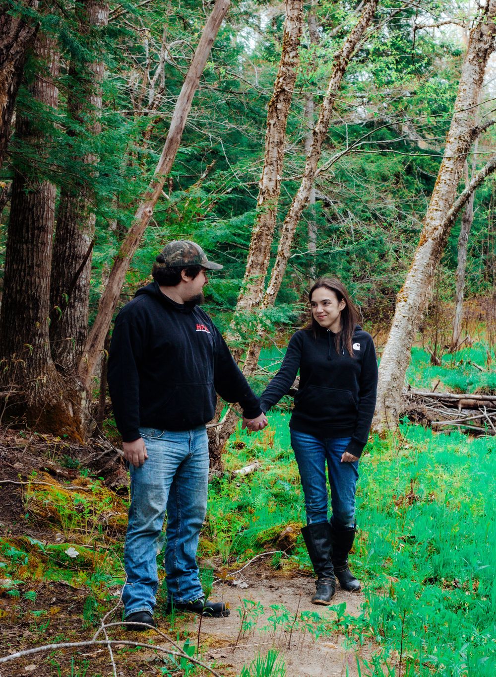 A couple holding hands while walking along a forest trail surrounded by evergreen trees.