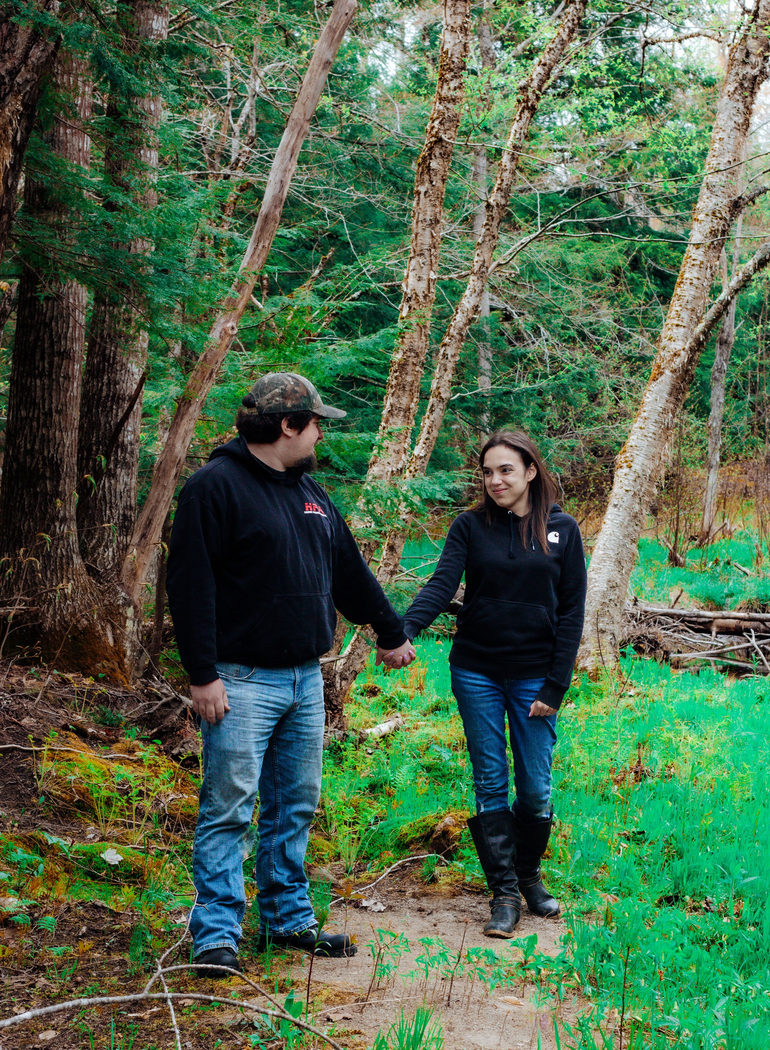 A couple holding hands while walking along a forest trail surrounded by evergreen trees.