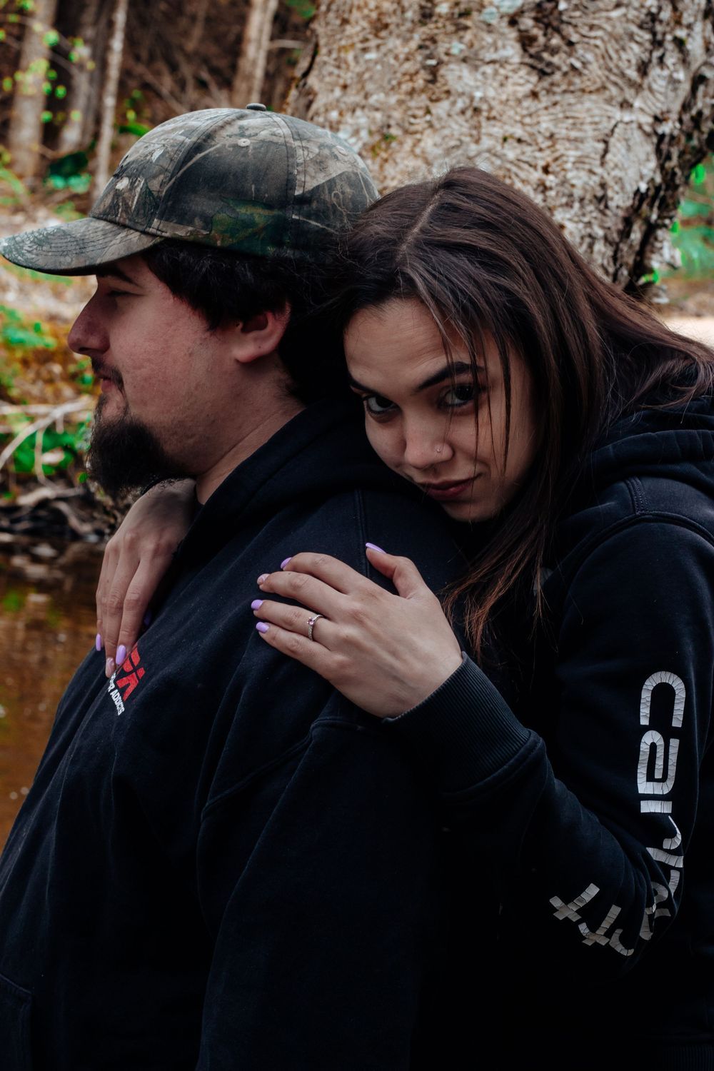 A couple in dark hoodies sharing an intimate moment near a stream in the woods.