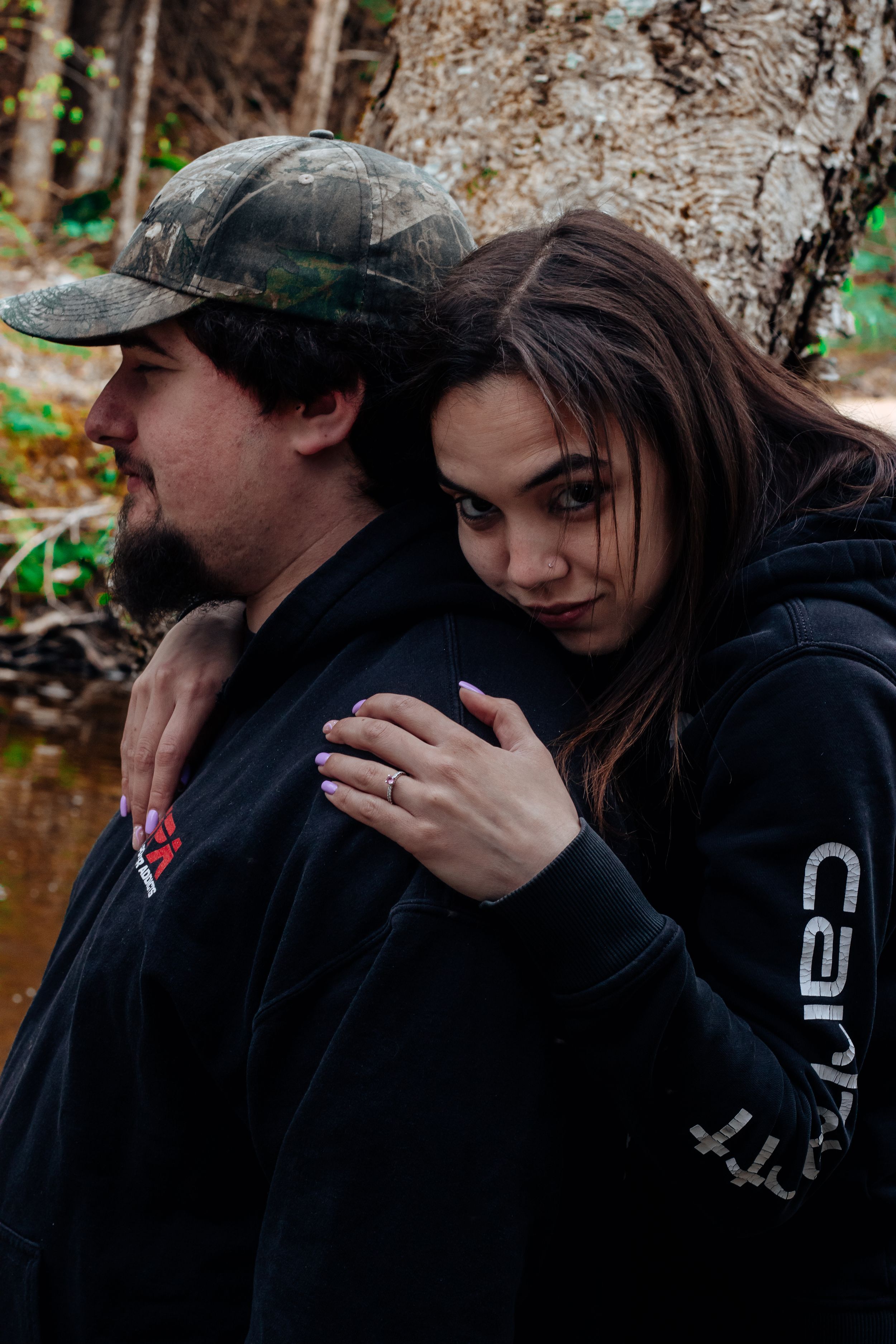 A couple in dark hoodies sharing an intimate moment near a stream in the woods.