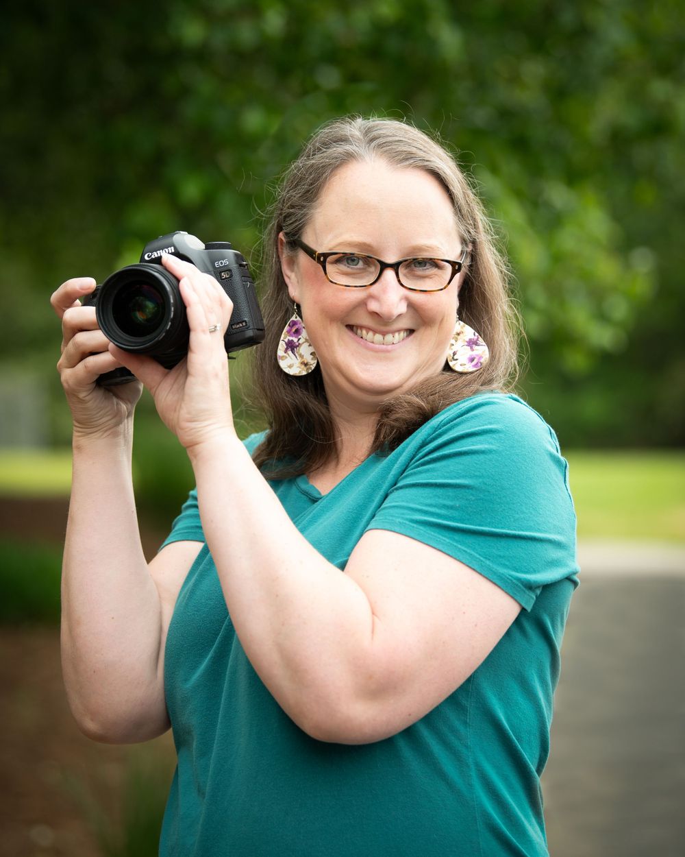 A smiling woman with glasses wearing a teal shirt holds a professional DSLR camera outdoors in a green setting.