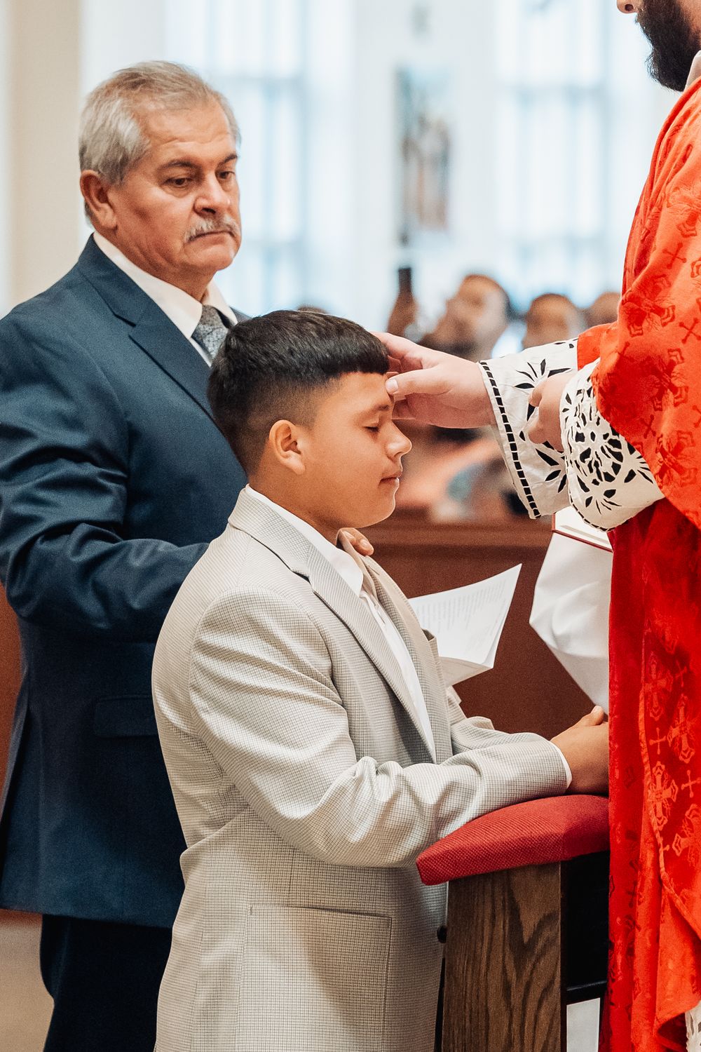 Young boy in suit receives blessing from priest during religious confirmation ceremony in church.