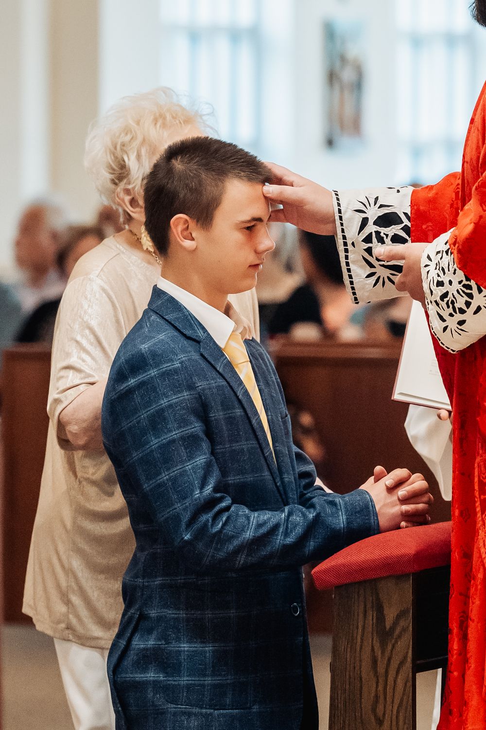 Young man in blue suit receives blessing from priest during confirmation ceremony in church.