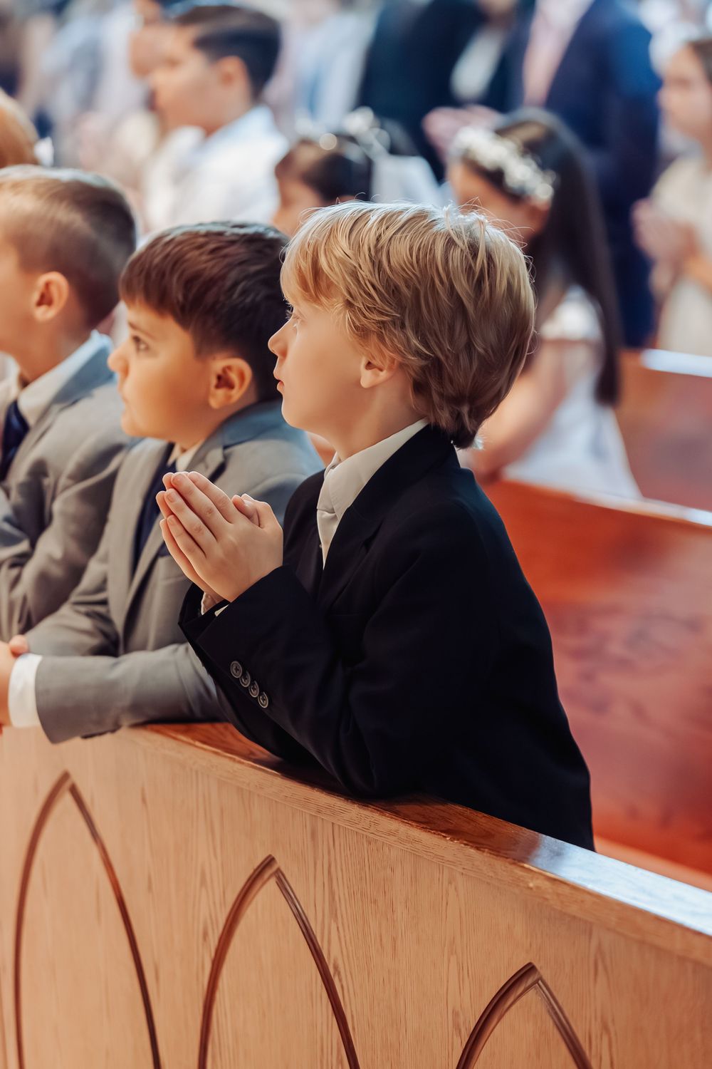 Boys in suits kneel in prayer at church pews during First Communion ceremony.