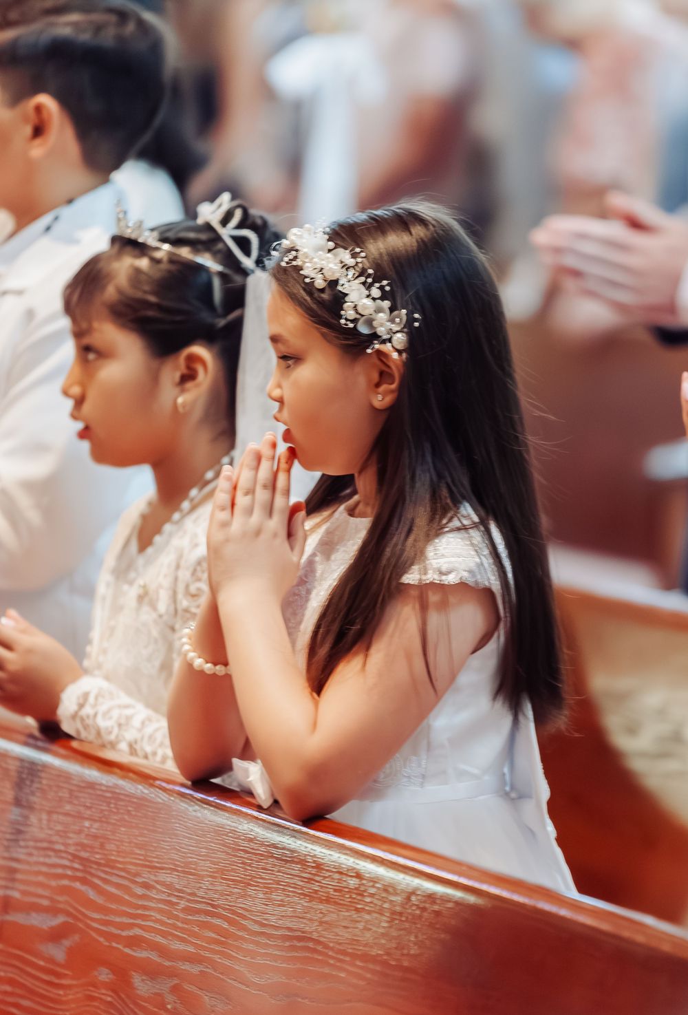 Young girls in white dresses and floral headbands pray reverently in church pews during First Holy Communion ceremony.
