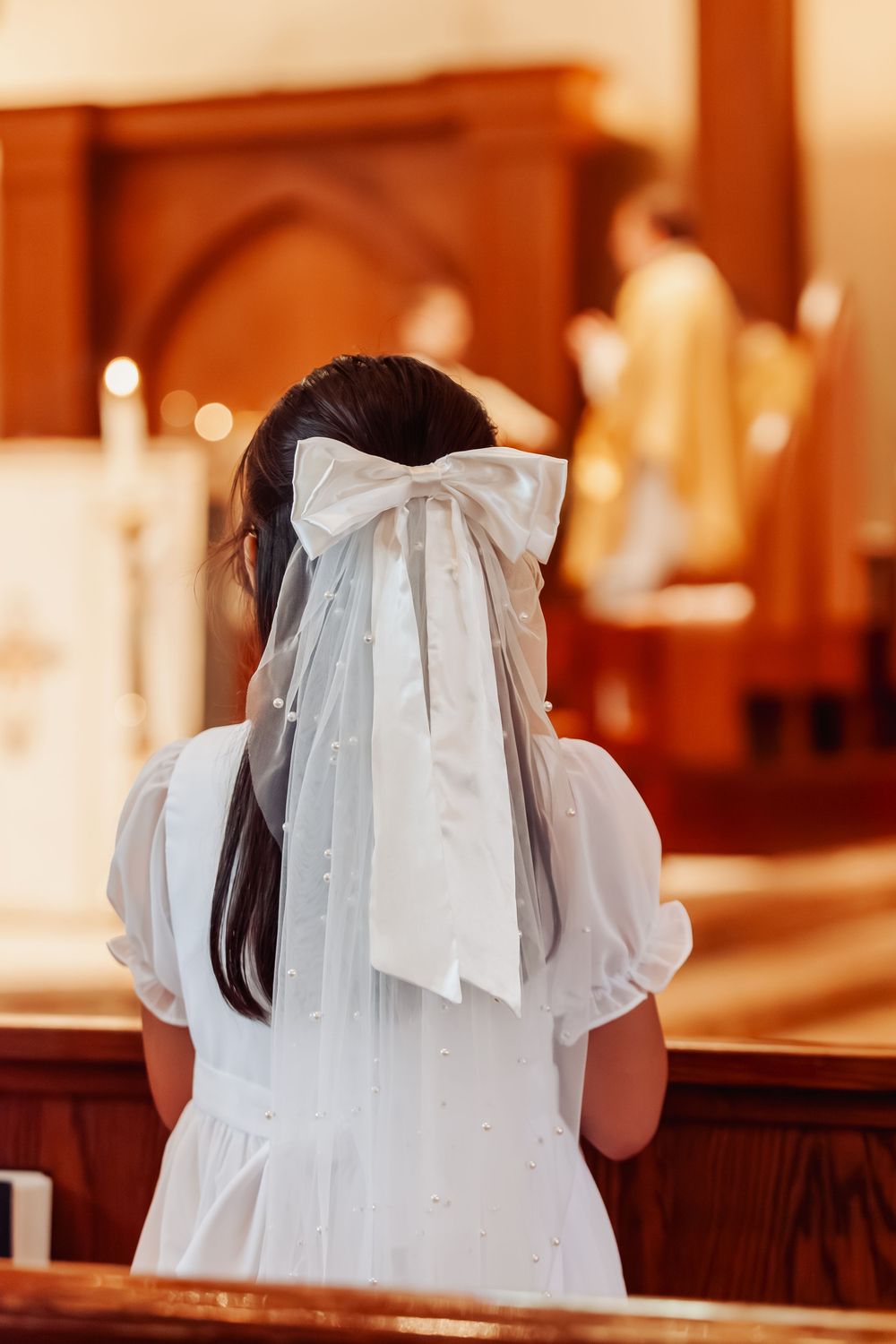 Girl in white dress and veil bow kneels at church pew during First Communion.