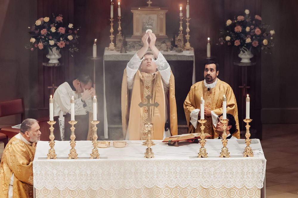 Catholic priests perform a solemn Mass at an ornate altar adorned with golden candlesticks and floral arrangements.