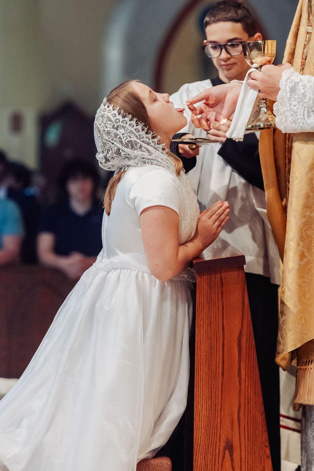 Young girl in white dress receives First Communion at church altar, kneeling with hands folded in prayer.