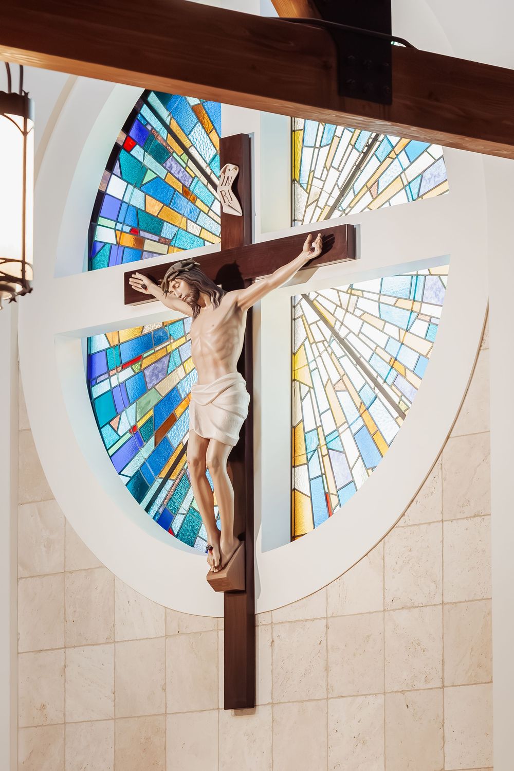 Crucifix statue of Jesus Christ on wooden cross with colorful stained glass window in modern Catholic church.