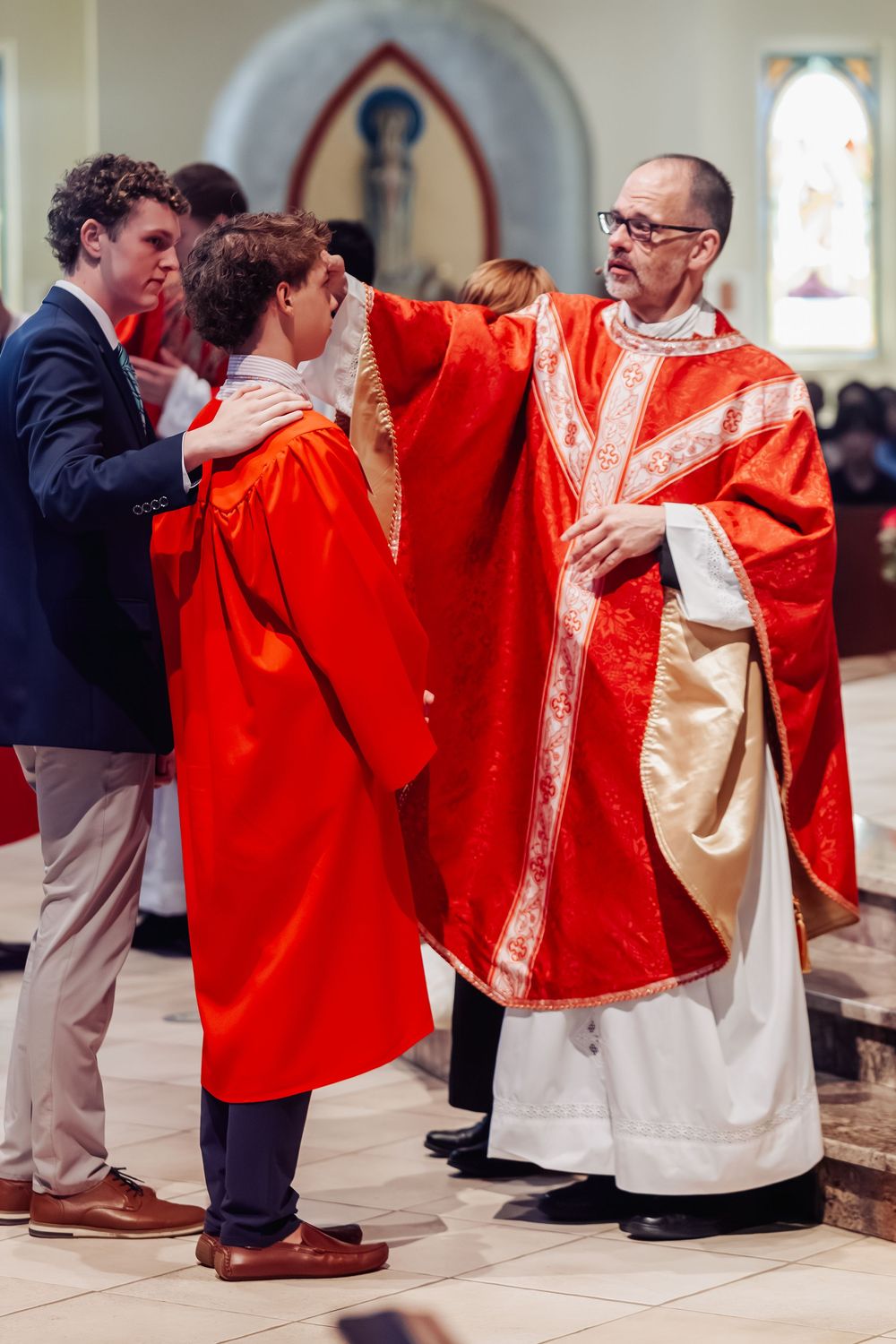 Priest in red vestments assists young person during a religious confirmation ceremony inside a church.