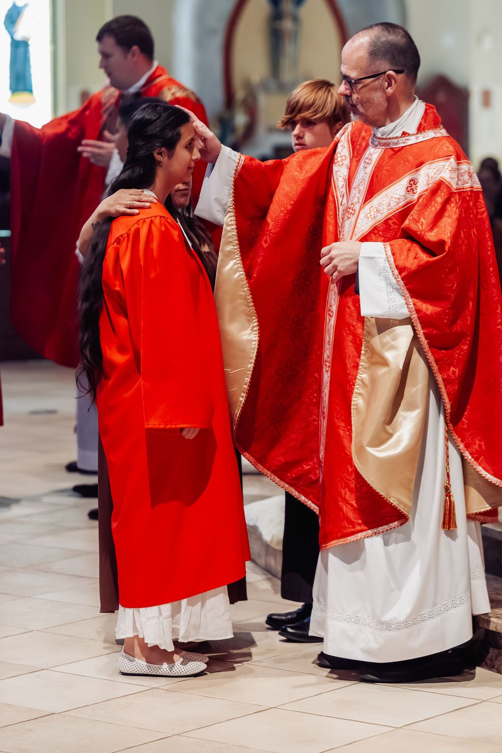 Bishop in red vestments performs Confirmation sacrament on young girl in red robe during Catholic church ceremony.