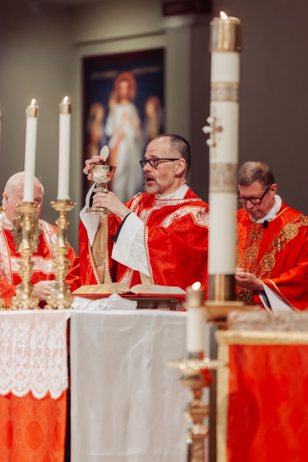 Catholic priest in red vestments elevates the Eucharist during Mass at a decorated altar with candles.