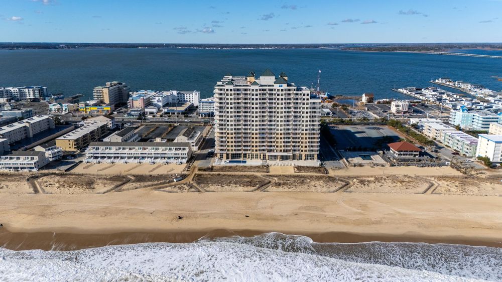 Aerial view of a beachfront high-rise condominium building on Ocean City with ocean waves and marina in background.
