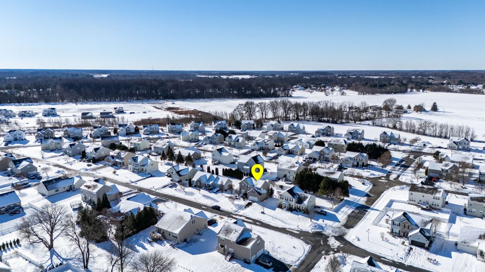 Aerial winter view of a snow-covered residential neighborhood with houses and streets stretching to a distant treeline.
