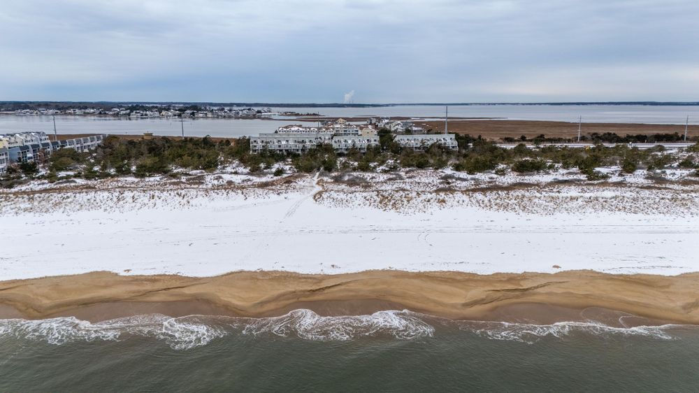 Aerial view of snow-covered beach dunes along coastal shoreline with winter waves crashing against sandy beach.