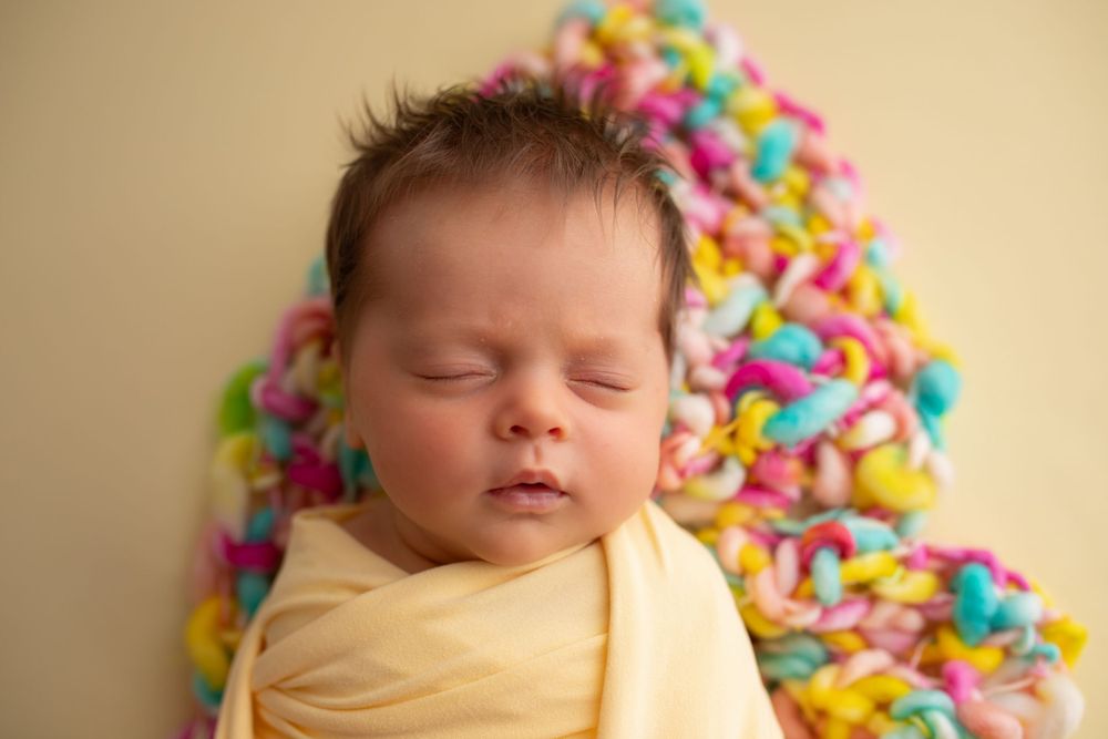 Newborn in yellow wrap against colorful rainbow yarn backdrop.