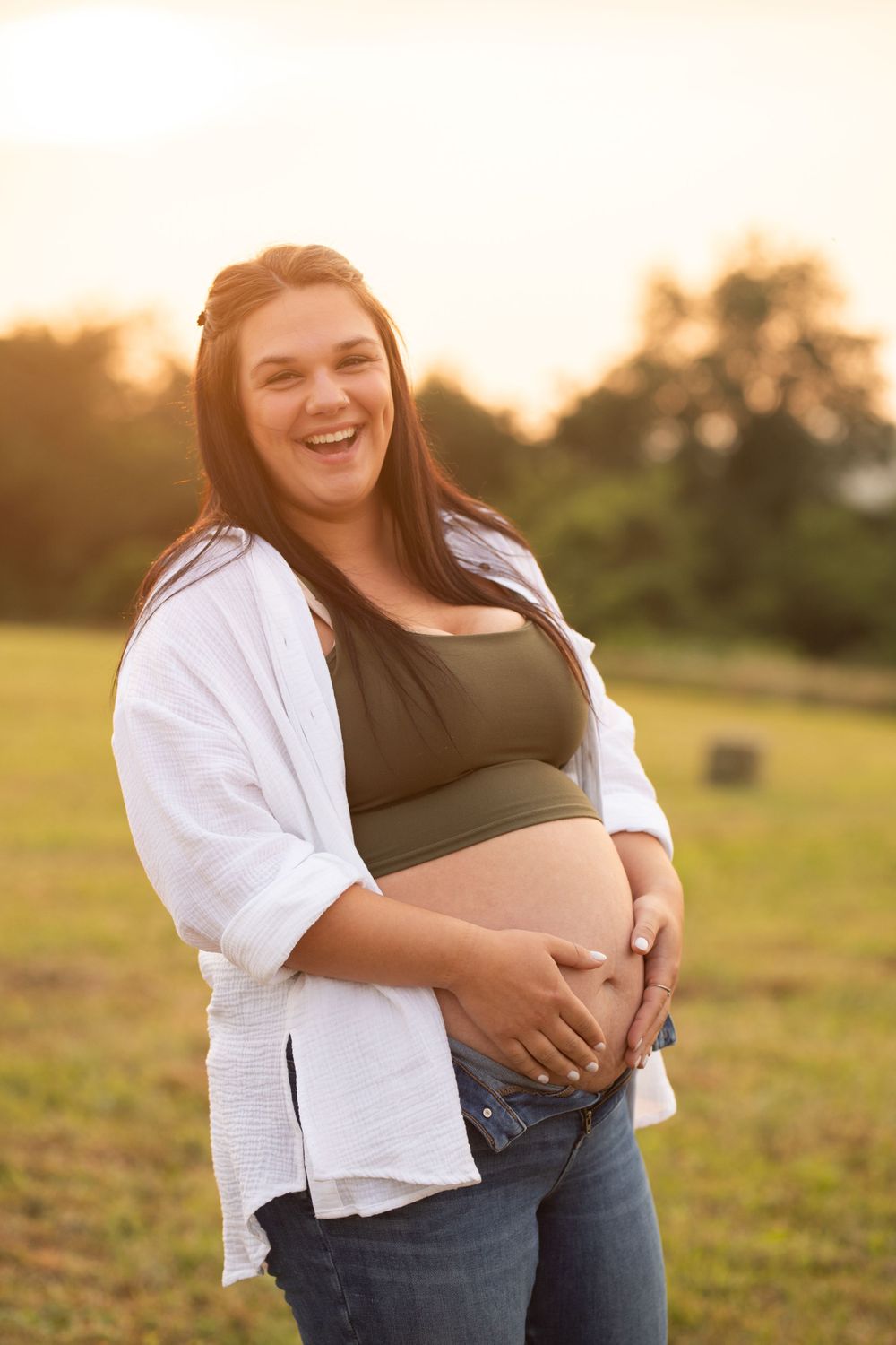 Expecting mother in white shirt and jeans poses joyfully in a golden sunset field.